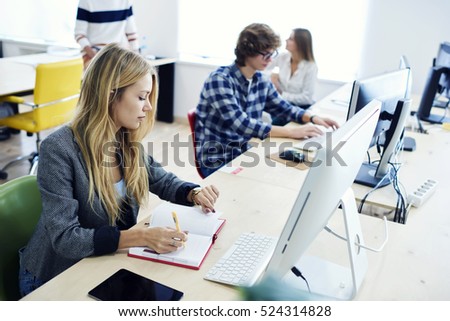 Young female student finishing individual university exam testing using special educational program and software via computers, looking at watch while writing down correct answers into notebook