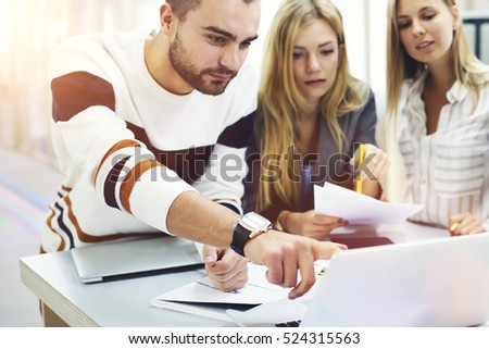On frontage, professional media coach pointing mistakes in students pres-release published on web page while on blurred background two young female pupils listening explanation making notes with pen
