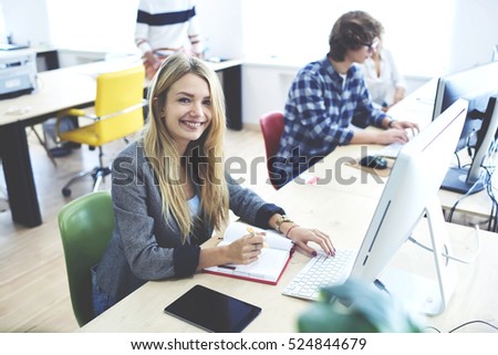 Young student in good mood working on exam tasks using modern computer and fast wireless connection to internet making notes of data information research during lesson in university coworking space