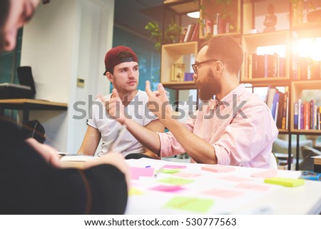 Two male skilled team leaders talking together about innovation in programming software for architect, discussing functionality of applications while sitting at the table in modern coworking office