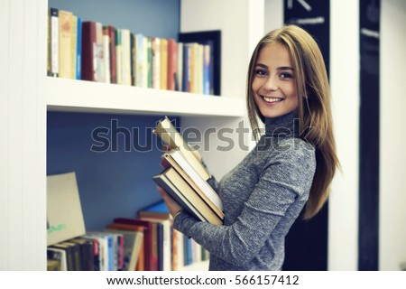 Cheerful female international student putting in order books returned after reading for literature lesson standing near bookshelves in modern interior library of university during break between lesson