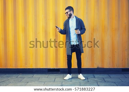 Handsome bearded male hipster making photo for blog sharing positive emotions feeling excited by sunny morning while walking to work with cup of coffee using modern smartphone and 4G internet