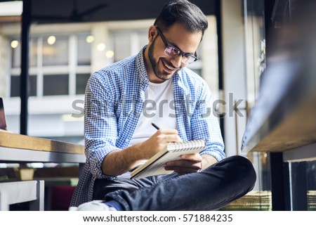 Handsome male freelancer writer creating advertising article for marketing company noting best ideas into diary before finishing publication fro release working hard in morning sitting in cafe