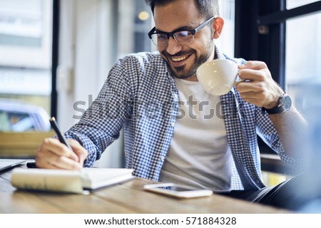 Cheerful male journalist in trendy glasses happy to finishing work on book review rereading written in notebook text before calling to editor to present publication drinking coffee for lunch in cafe
