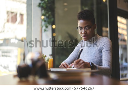 Experienced female editor of fashion magazine working on creation of book review writing  best ideas into notebook before publishing on web page of issue online version sitting in modern cafe