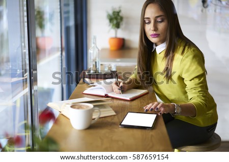 Young attractive female administrative manager working with financial documentation and financial reports in online database checking accountings via touchpad with mock up screen connected to wifi