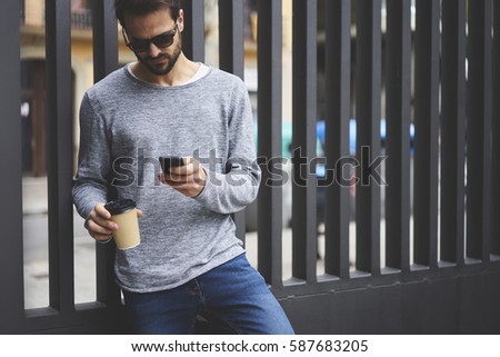 Handsome bearded guy strolling on city street with coffee to go using online navigator to find right direction using modern smartphone connected to fast 5G wireless in roaming enjoying weekends