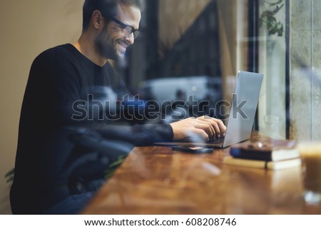 Smiling bearded entrepreneur chatting with friends in social networks and watching funny video online using modern technology and free internet connection while sitting in cozy coffee shop interior