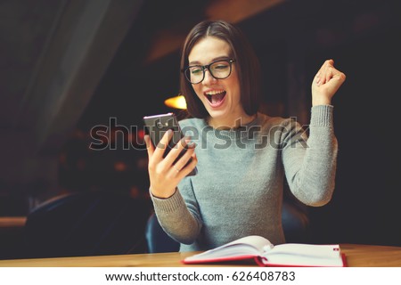 Emotional young attractive female expressing surprise checking email box and reading messages with congratulation of winning using modern smartphone connected to 4G internet in coworking office