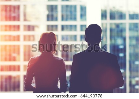 Back view silhouettes of two business partners looking thoughtfully out of a office window in situation of bankruptcy,team of businesspeople in fear or risk watching cityscape from skyscraper interior
