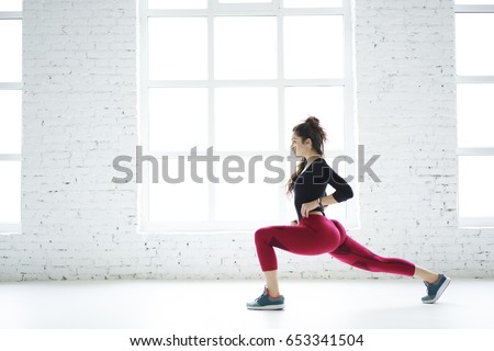 Young slim sportswoman doing squat exercises working on lower body legs and butt muscles create perfect body shape, determinate athletic girl tensing muscles during training set in white modern gym