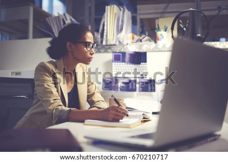 Experienced female administrative manager in trendy eyewear organizing financial documents for upcoming business meeting with executive using modern computer and wireless internet in office indoor