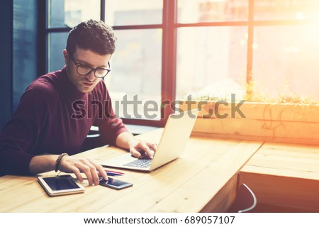 Concentrated professional IT developer in stylish eyeglasses checking notification on smartphone while creating new web application on laptop computer connected to 4G during remotely job indoors