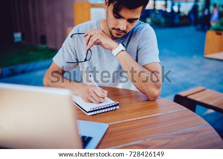 Pensive male student making plan of preparation for exams writing goals in notepad while sitting on terrace of cafe, concentrated hipster guy making notes while making researches for startup on laptop