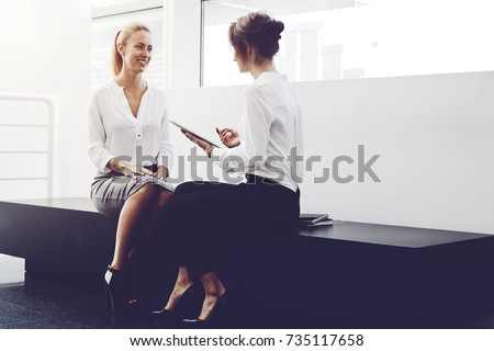 Young female lawyer is holding touch pad and talking something to her smiling client, while they are sitting in modern office interior. Smart woman using digital tablet for consultation with customer