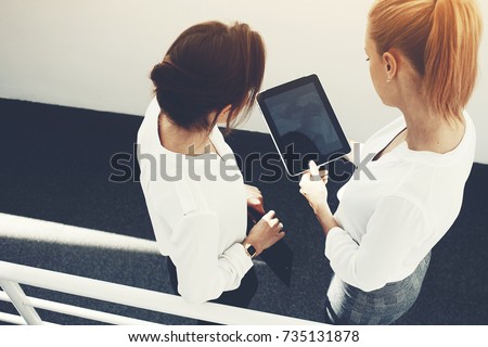Top view of woman CEO is holding touch pad with mock up copy space on the screen, while is standing with client in corridor company. Two female are watching video on digital tablet before conference