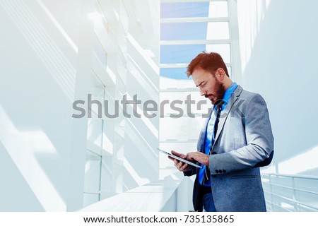 Portrait of young prosperous business man dressed in suit work on digital tablet during break, skilled male entrepreneur dressed in corporate clothes using touch pad while standing in modern office