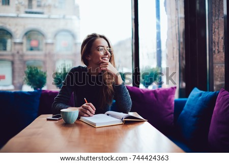 Positive young woman in stylish eyeglasses dreaming while sitting at table with opened book in stylish coworking space.Smiling beautiful female in spectacles looking away during reading bestseller
