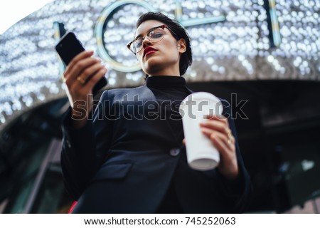 Serious female entrepreneur with red lips updating application on mobile while strolling on street, confident businesswoman in formal wear making money transaction online on coffee break outdoors
