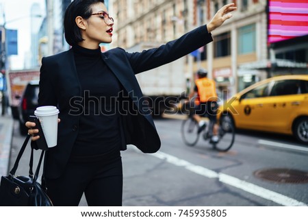 Attractive businesswoman with red lips raising had to call yellow cab on New york avenue, confident trendy dressed female manager hailing on road catching taxi holding coffee to go getting to work