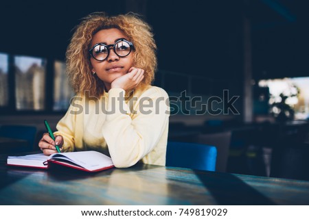 Thoughtful young woman in eyeglasses writing to do list of goals writing in diary in coffee shop, female student dreaming about weekends while making notes in personal organizer planning day