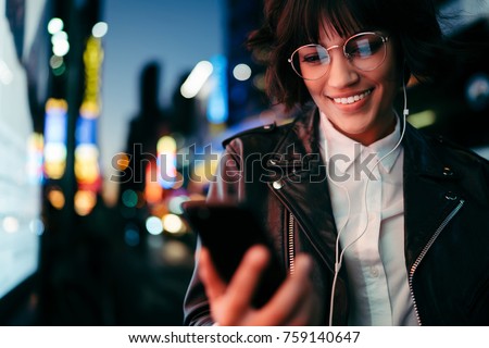 Smiling gorgeous girl browsing web site with music for downloading having fun on night city street with illumination, cheerful female hipster reading message on mobile listening songs from playlist