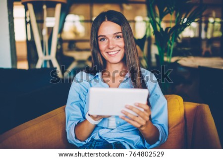 Portrait of cheerful young woman using portable pc for blogging in social networks sitting in coffee shop,happy hipster girl looking at camera while downloading new application from networks on tablet
