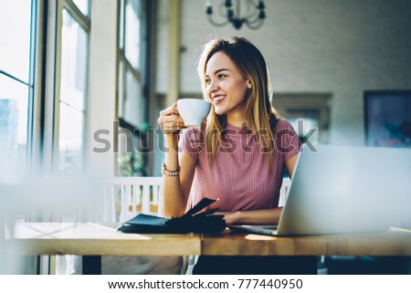 Cheerful cute hipster girl with blonde hair enjoying breakfast in cafeteria starting day with coffee.Female student resting in cafe with cozy atmosphere sitting at table with notepad and netbook
