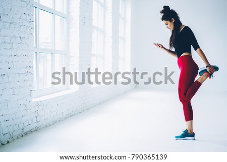 Young woman training in gym watch video from web page online on mobile for stretching muscles of lower body,sportswoman warming up and blogging in networks on smartphone standing near publicity area