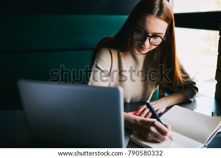 Skilled attractive student in trendy optical spectacles watching webinar on laptop device connecting to wireless internet and writing notes in notebook with mock op area for your advertising content