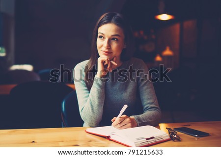 Pondering attractive young woman with pen in hand thinking on plans and writing list to do in notepad enjoying recreation time in stylish coworking space.Thoughtful student noting text information