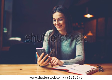 Positive female blogger reading comments under post in social networks on modern telephone device sitting in coworking space at wooden table with book.Copy space area for your advertising text