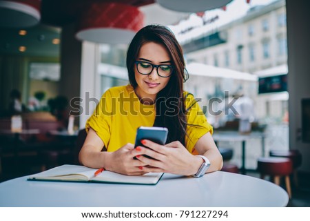 Young woman blogger in spectacles publishing new post on own website and installing new application for editing photos in social networks on digital smartphone device connected to free 4G internet