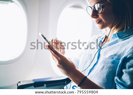 Young female meloman listening favourite songs during flight in first class cabin using mobile playlist and accessory, woman entertaining on airplane board enjoying music in headphones from smartphone