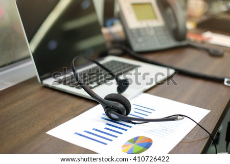 close up blurred of headset of call center hotline at computer office room:telemarketing business financial concept:headphone laptop telephone and statistics graph in image picture:telework corporate