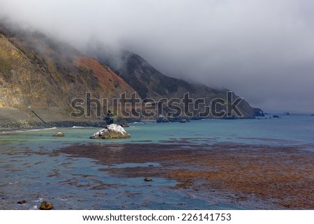 Cloud covered mountains rising from the ocean. Dark clouds in the distance, bright colors in the foreground, along with an offshore rock that is covered with bright white bird droppings.
