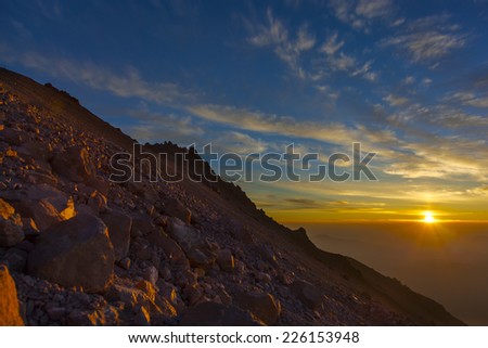 View into a rising sun from above the clouds and just below a mountain peak.