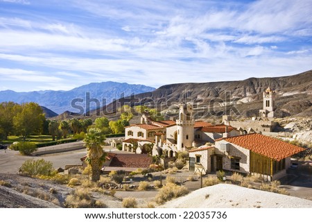 Spanish villa between mountains. Multiple buildings with ceramic roof tiles and wooden shutters on the windows. California fan palms near the villa. Mountains of sedimentary rock in the background.