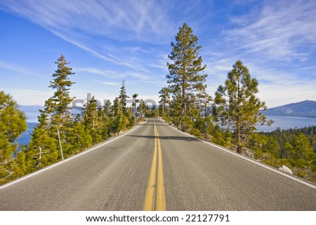 A two lane highway high above an alpine lake, yellow divider line in center of image. Pine trees on both sides of the highway. Mountains in the distance. Blue sky with cirrus clouds.  Lake Tahoe.