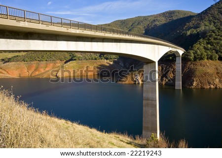 A concrete bridge spans a reservoir. Vegetation free slopes above the water indicate its level is below full capacity. Dense forest on the hills above the reservoir. Blue Sky with thin clouds.
