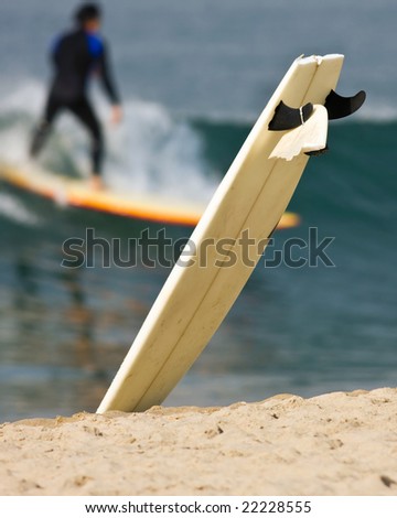 A damaged surfboard stuck in the sand with one end up in the air. Its broken-off tail is tied to one of its fins. Out of focus surfer and wave in the background.