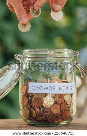Hands putting coins in a money jar full of coins with the word crowdfunding on it.