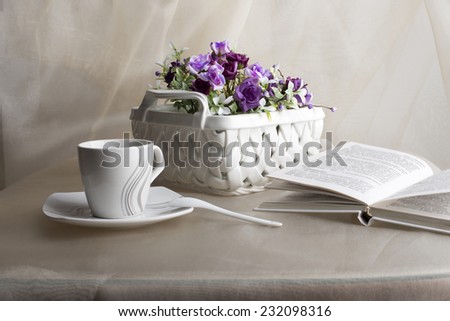 White teacup, teaspoon, vase with violet flowers and book on the beige organza