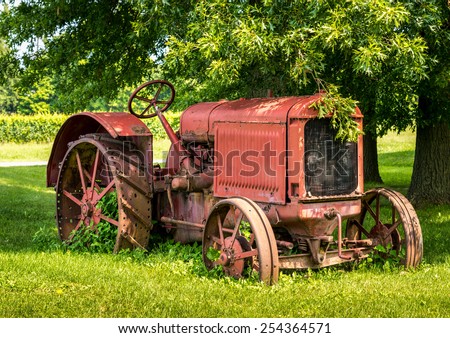A old red tractor sits under a tree on the edge of a farm field.