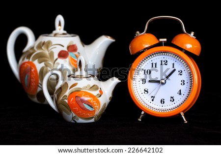 Orange alarm clock and decorative tea pots beside on black velvet texture background.  Tea time. Colorful lifestyle. Modern stylish design