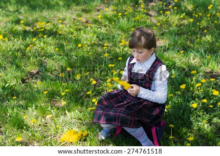 little girl in a white blouse and school sundress in a cage in the park sitting on the portfolio. kid weaves a wreath of dandelions