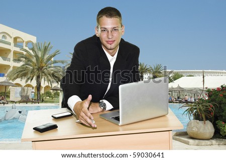 Young businessman holding hand over his desk with laptop, mouse and mobile, touristic resort with pool and hotels in the background