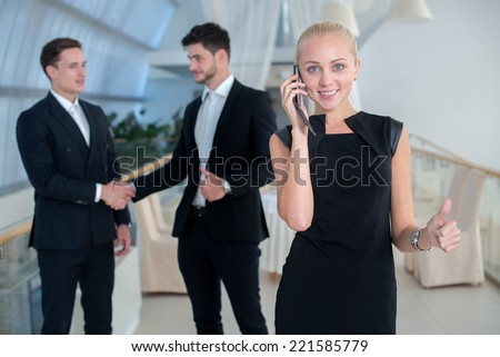 Talking by mobile phone. Successful business woman speaks by mobile phone. Two businessmen are standing on the background and shaking their hands