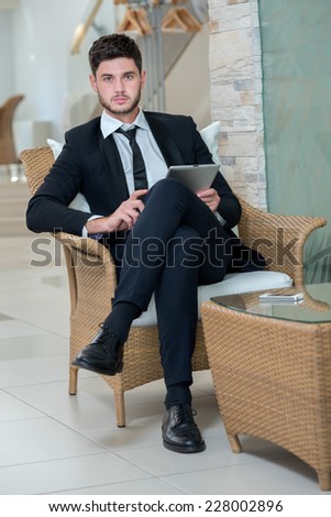 Confident and successful businessman. Young and motivated businessman is sitting in the chair, working with tablet and confidently looks forward