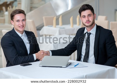 Well done. Tho stylish and motivated businessmen are shaking hands looking directly in the camera.